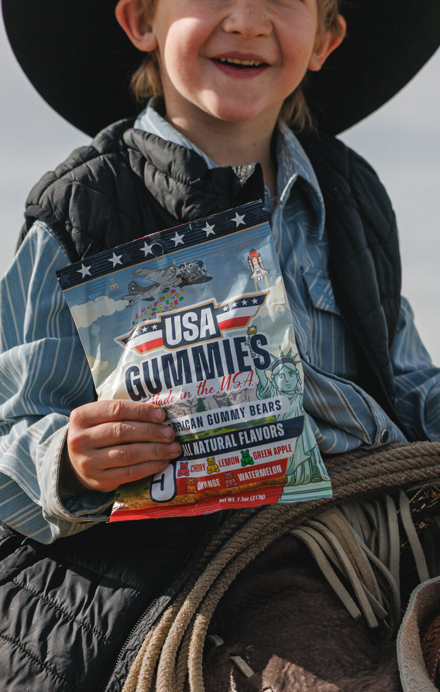 A smiling young cowboy on horseback holding a bag of USA Gummies, mountains behind him