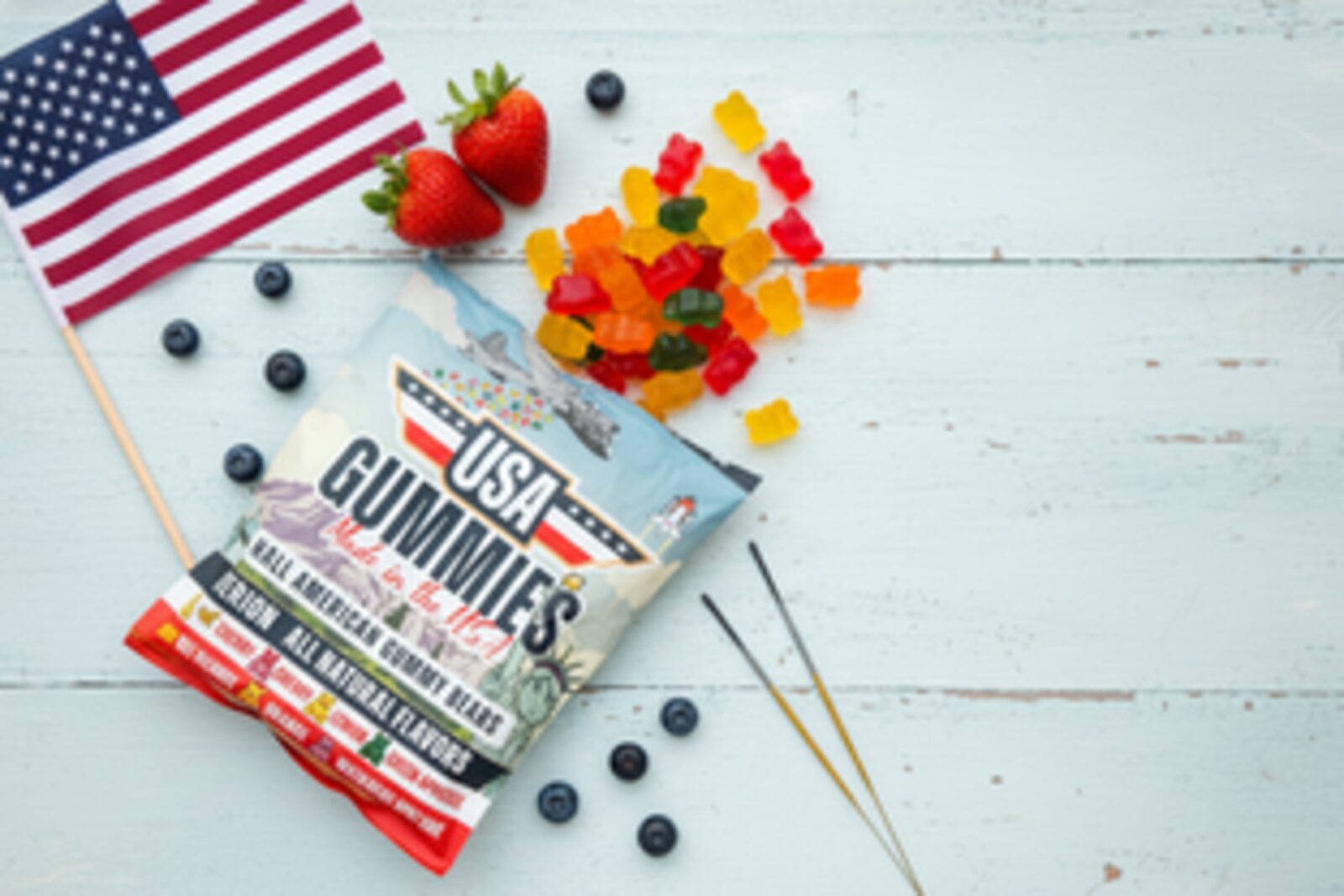 An American flag, a bag of USA Gummies, real strawberries and blueberries, and gummy bears arranged on a light wood surface